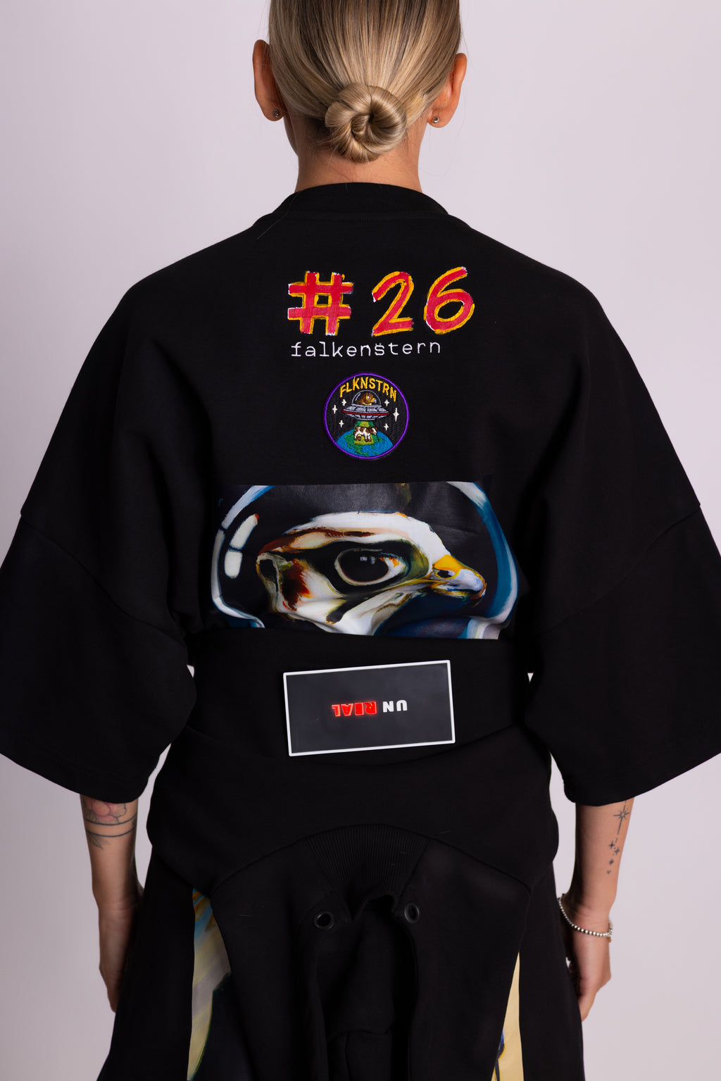 Person in einem schwarzen T-Shirt mit Grafikdesign und Text auf dem Rücken vor dunklem Hintergrund