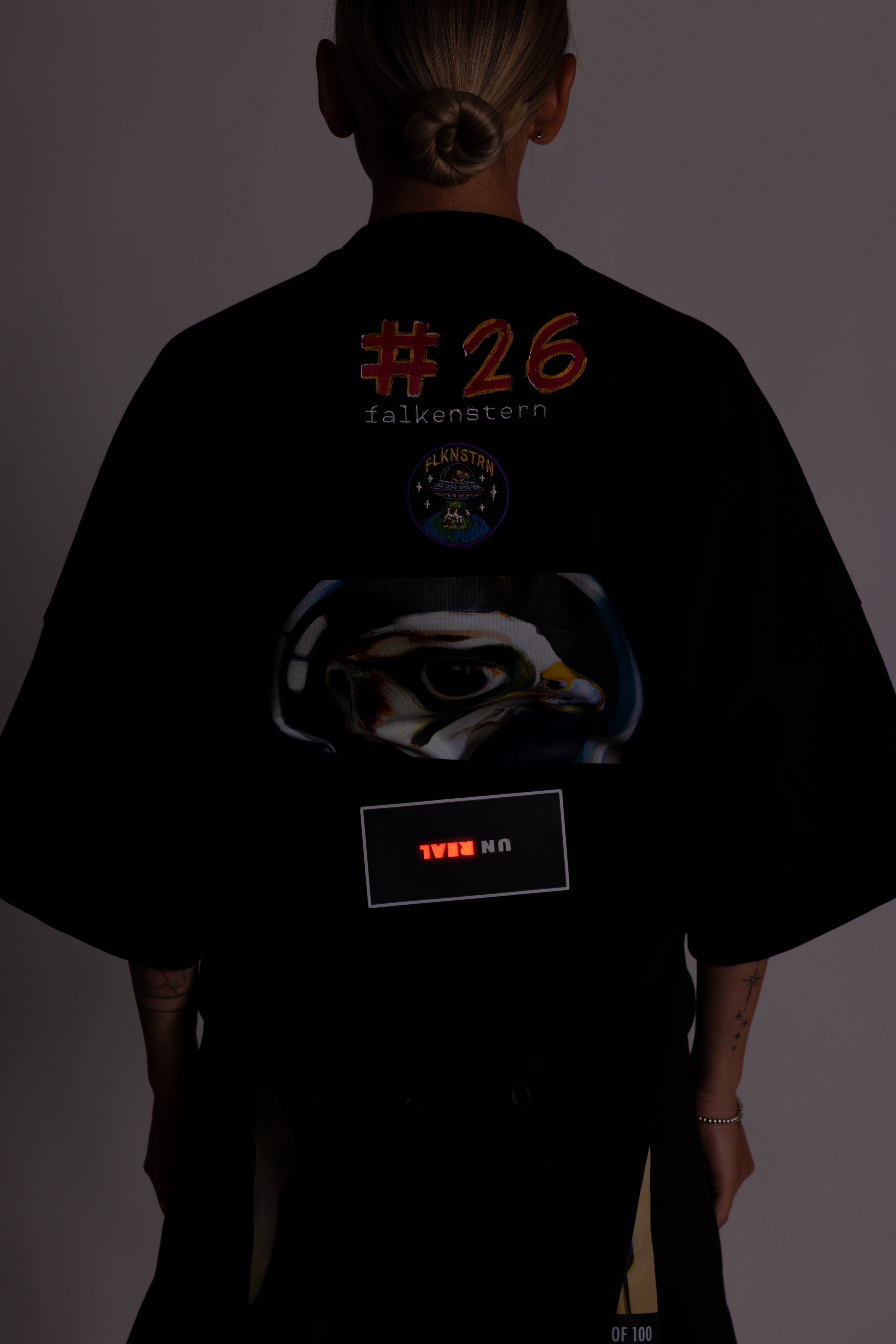 Person in einem schwarzen T-Shirt mit Grafikdesign und Text auf dem Rücken vor dunklem Hintergrund