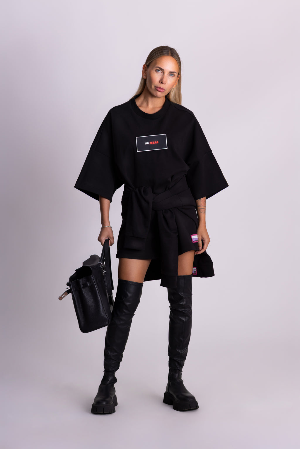 Person in schwarzer Kleidung mit Logo, die eine schwarze Tasche hält, vor hellgrauem Hintergrund