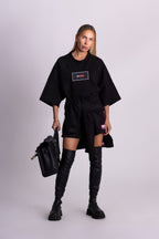 Person in schwarzer Kleidung mit Logo, die eine schwarze Tasche hält, vor hellgrauem Hintergrund