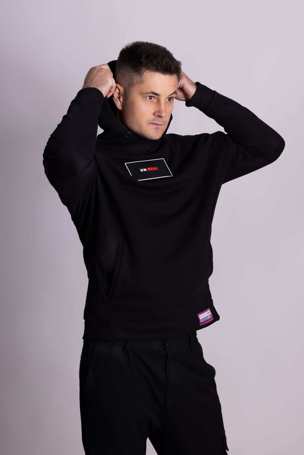 Mann in schwarzer Kleidung mit sichtbarem Markenlogo vor hellgrauem Hintergrund