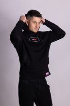 Mann in schwarzer Kleidung mit sichtbarem Markenlogo vor hellgrauem Hintergrund